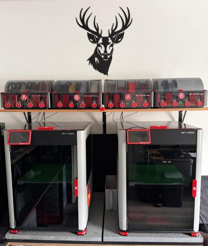 Zwei 3D-Drucker mit einem stilisierten Geweih darüber auf einer Arbeitsfläche.